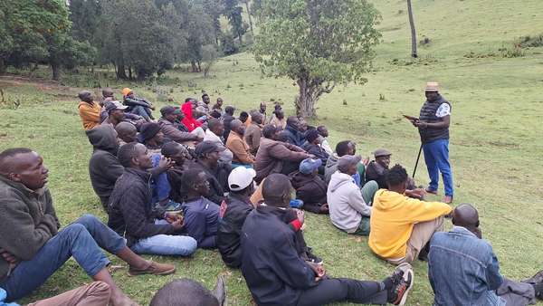 Residents seated on a grassy hillside listen to Long’ole