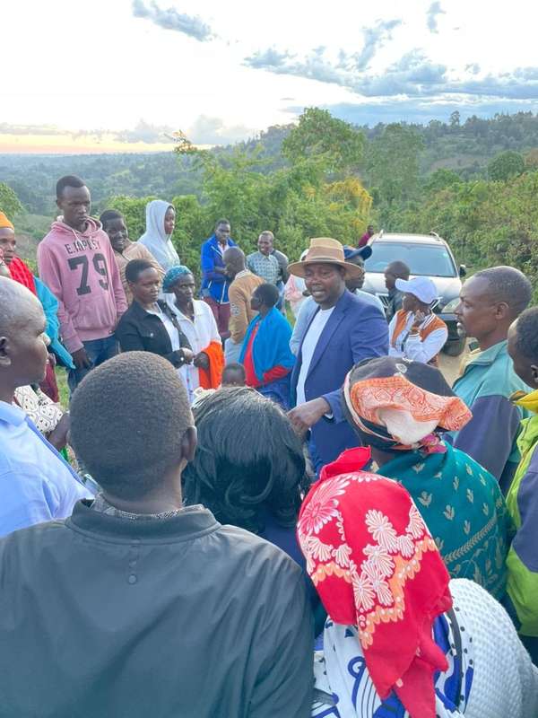 Long’ole talks with a crowd of residents at dusk, hills behind