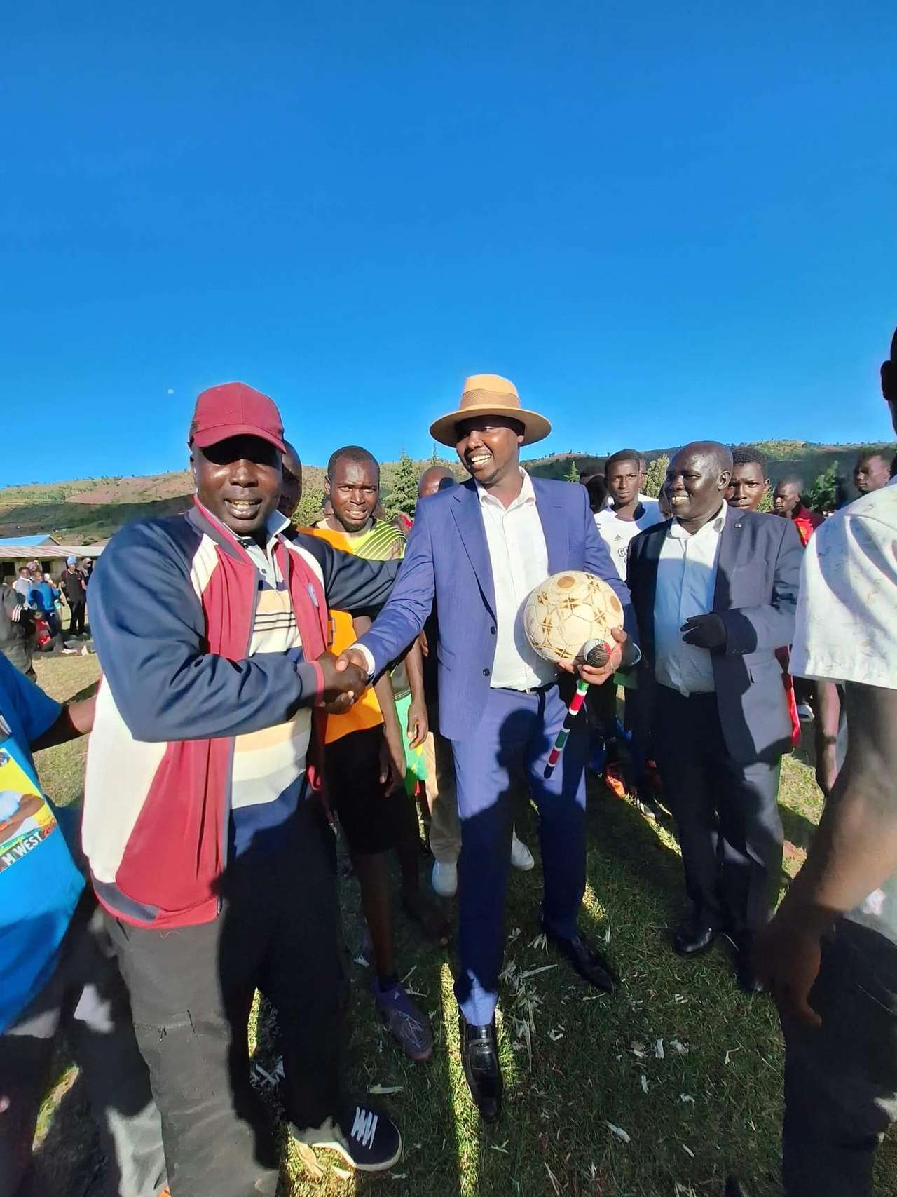 Long’ole, in a blue suit and hat, shakes hands with a supporter while holding a football, surrounded by young players