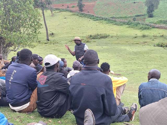 Long’ole addressing men seated on a grassy hillside above farmland