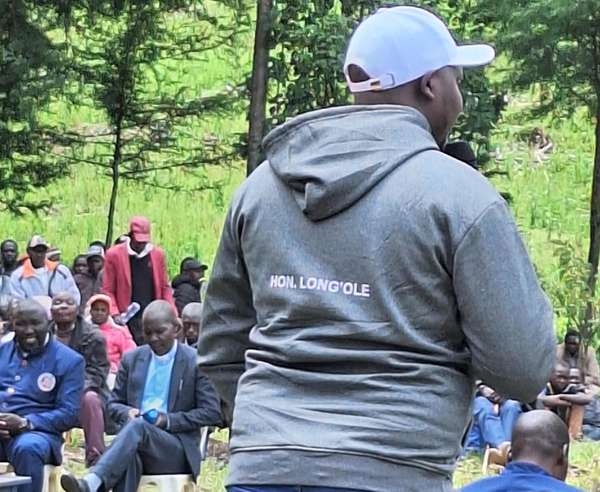 Long’ole, seen from behind in a grey hoodie printed HON. LONG’OLE, addresses seated residents