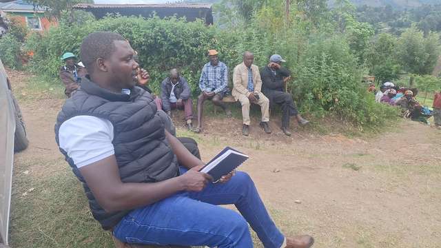 Long’ole seated with a notebook, listening to residents at an outdoor meeting