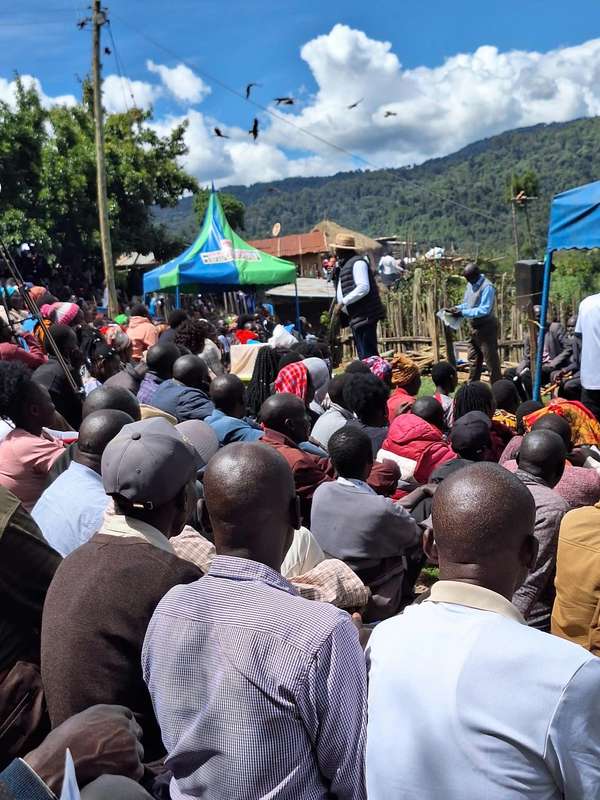 A large seated crowd listens to speakers under blue and green tents, forested hills behind